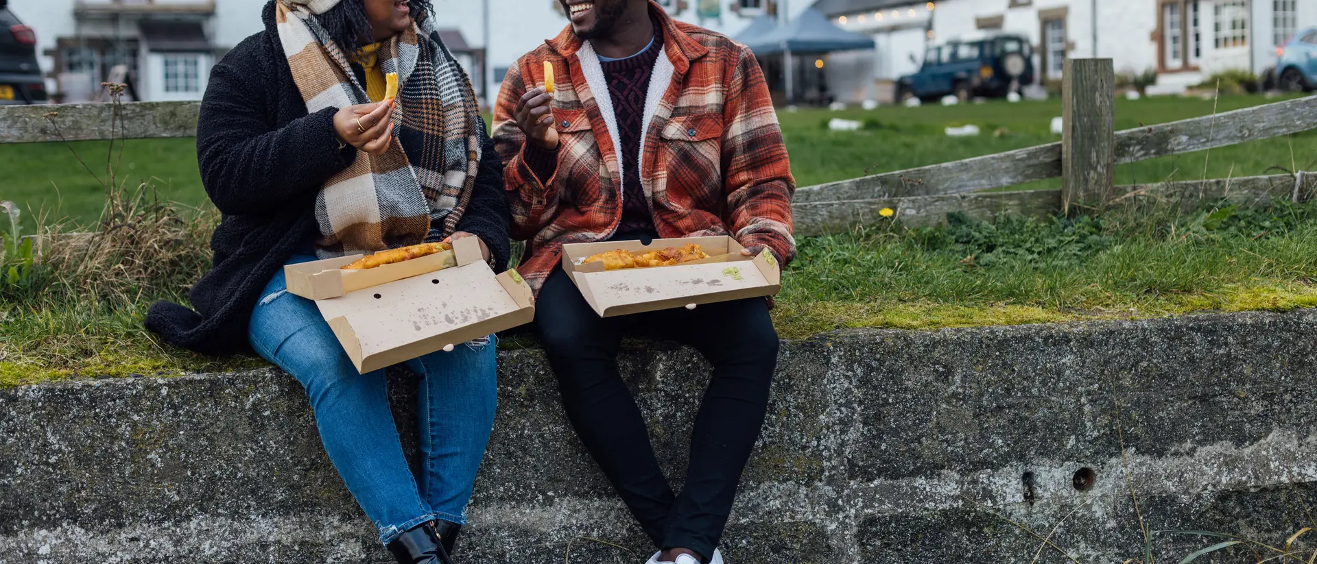 Two people enjoy food from takeout boxes while sitting on a stone wall, with buildings and greenery in the background.