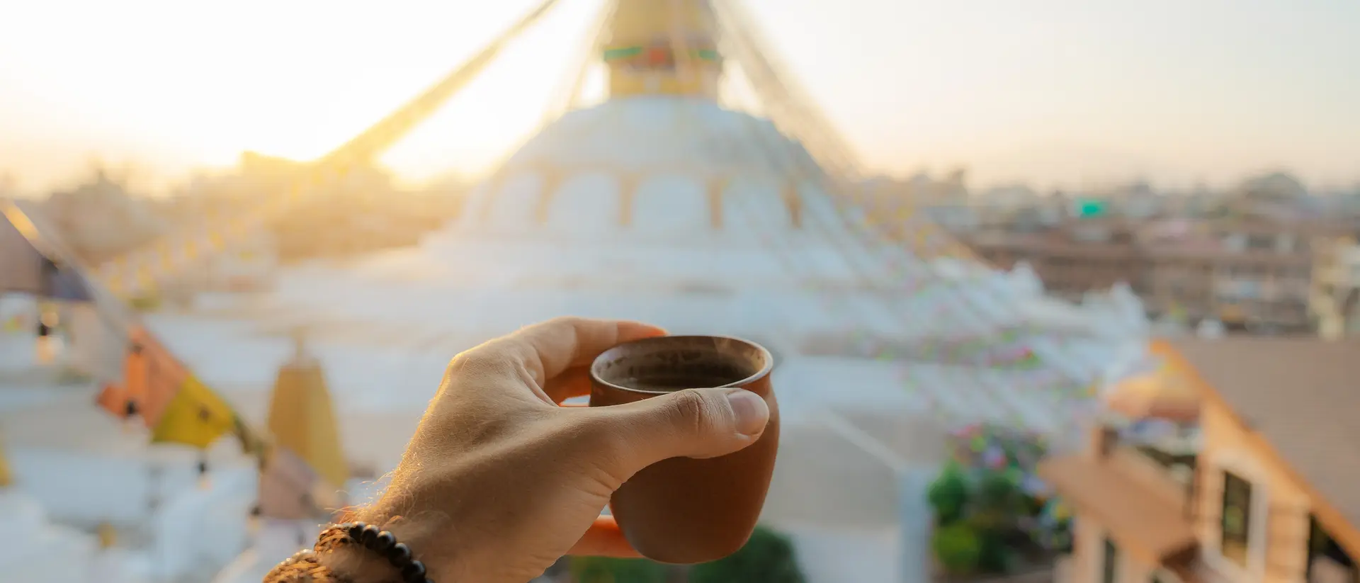 A hand holding a small clay cup in front of a stupa at sunrise, with colorful prayer flags in the background.