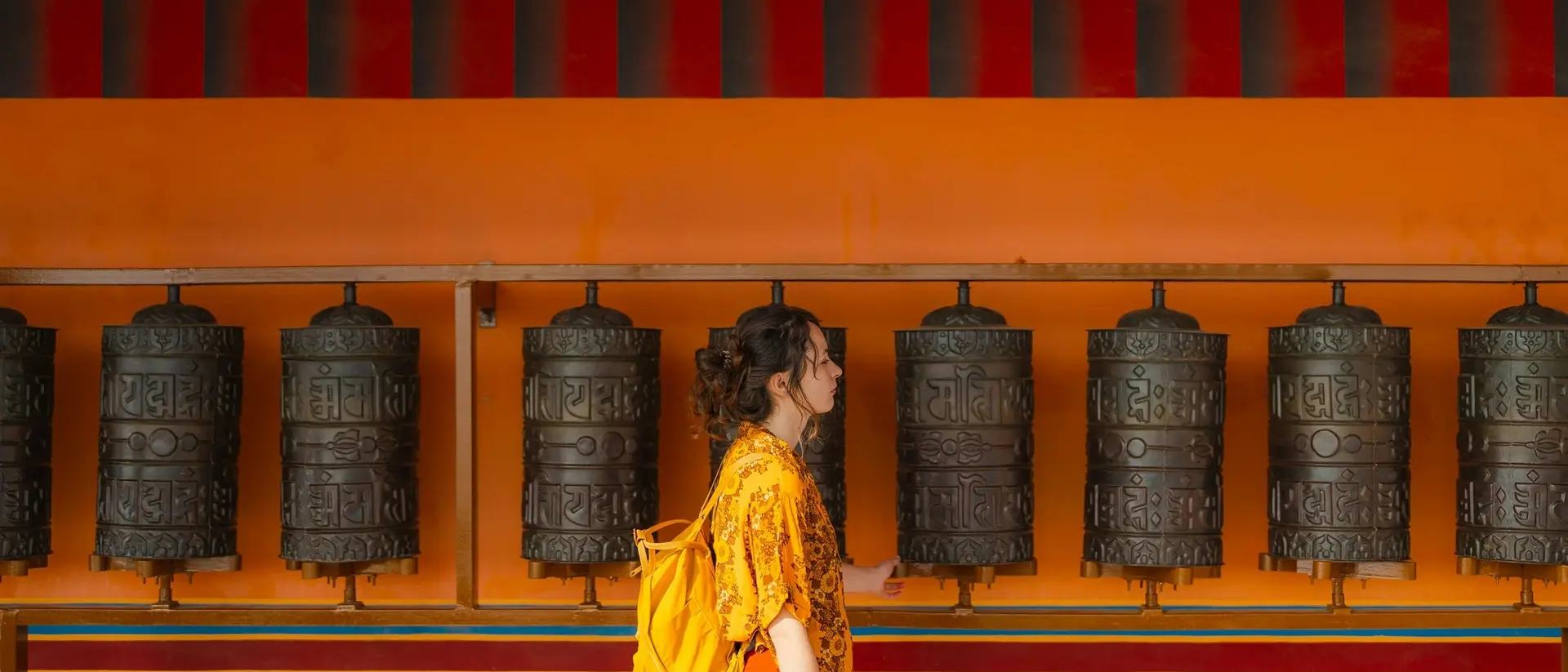 A woman in a yellow shirt and orange skirt walks past a row of prayer wheels against a vibrant orange and turquoise wall.