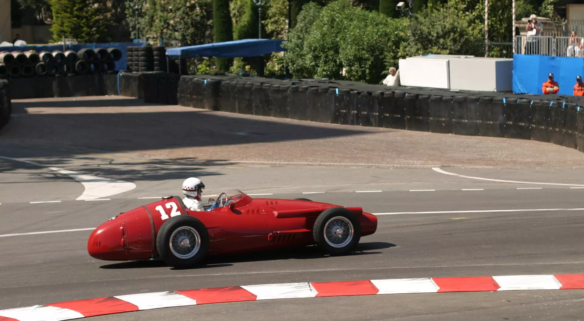 A vintage red race car, number 12, speeds around a bend on a racetrack with spectators in the background.