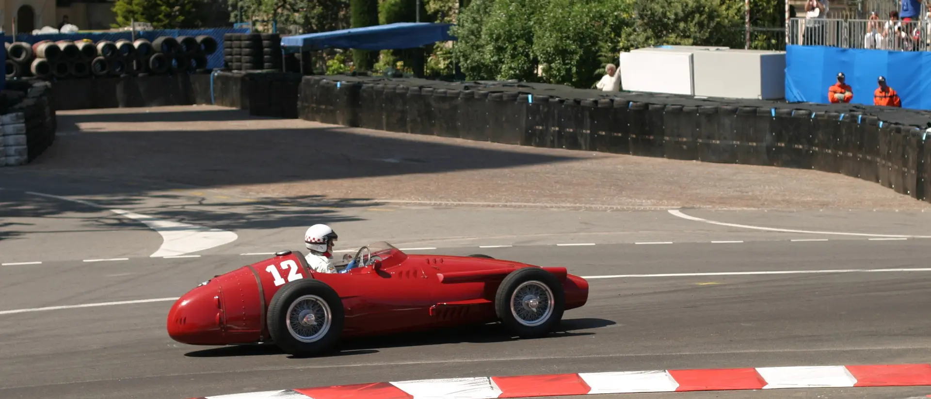 A vintage red race car, number 12, speeds around a bend on a racetrack with spectators in the background.