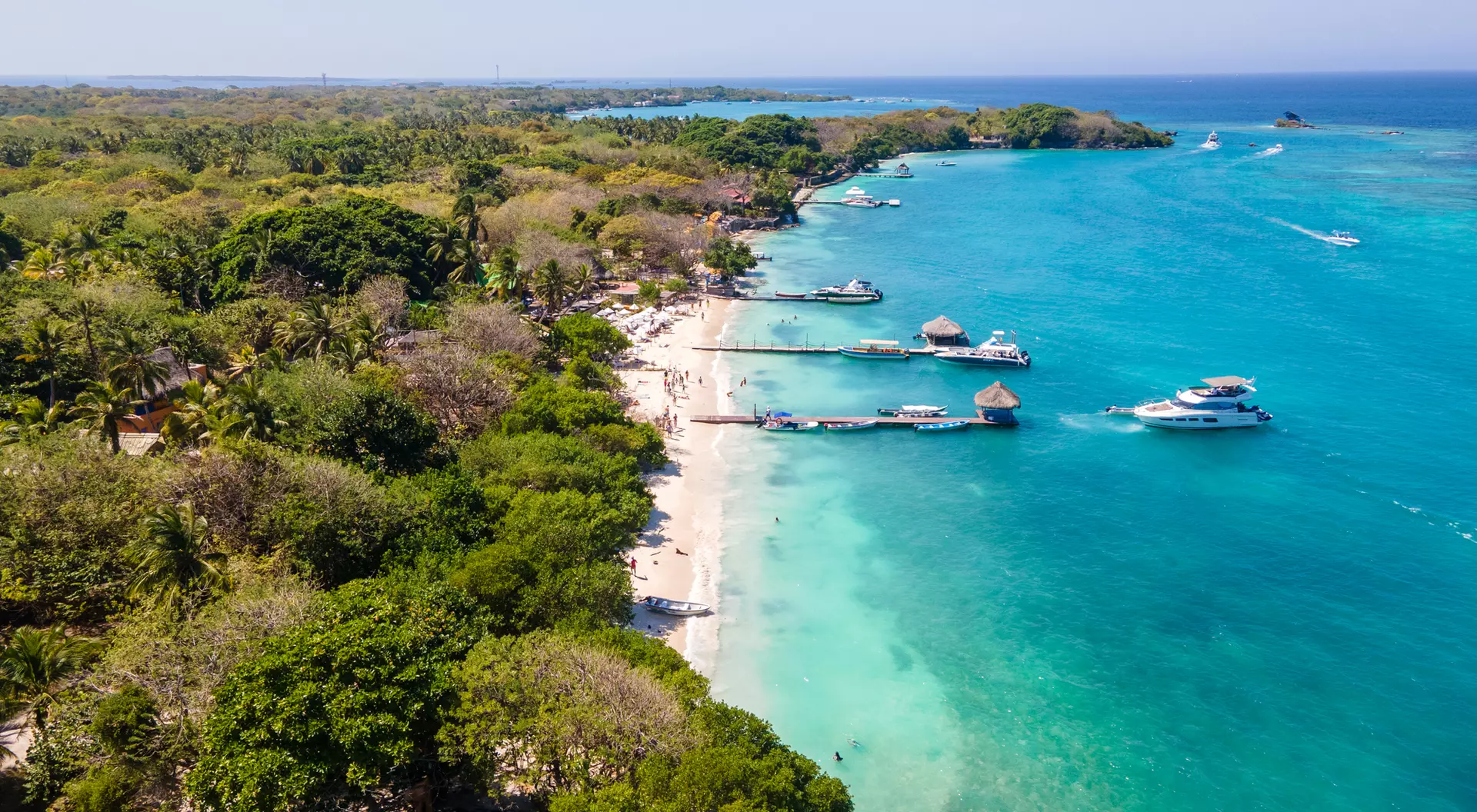 Aerial view of a tropical beach with clear turquoise water, boats, and lush green trees lining the shore.