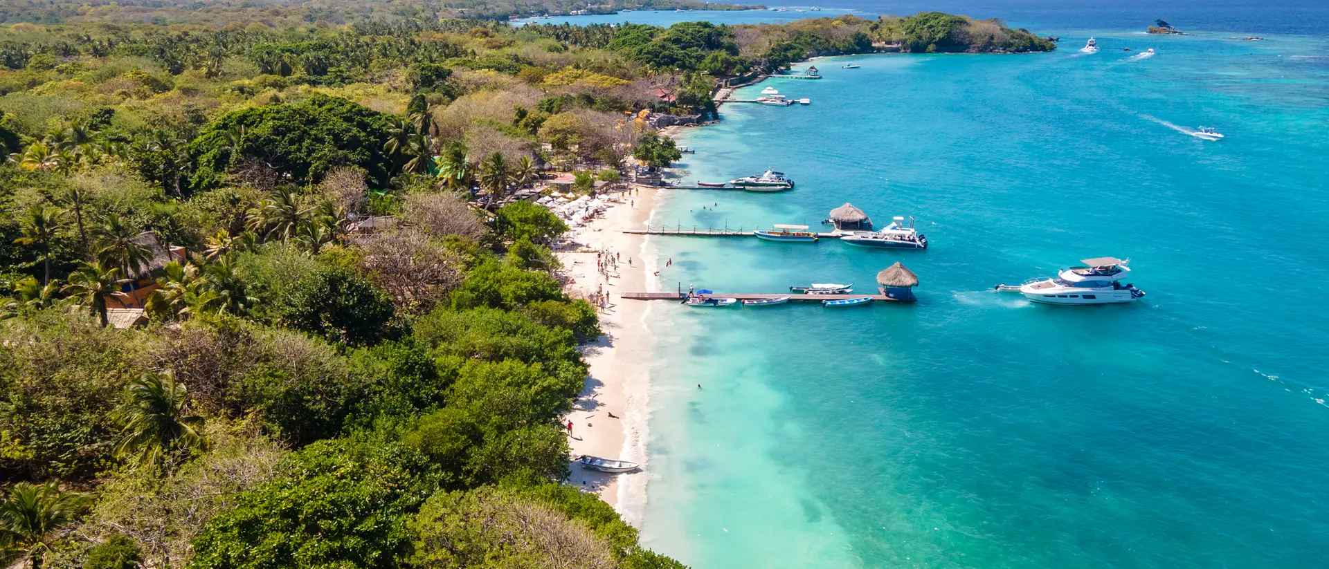 Aerial view of a tropical beach with clear turquoise water, boats, and lush green trees lining the shore.