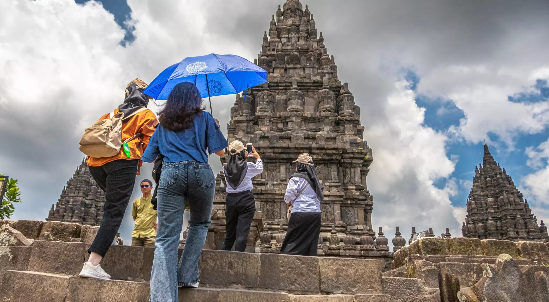 Visitors ascend stone steps toward ancient temples under a partly cloudy sky, some using umbrellas for shade.