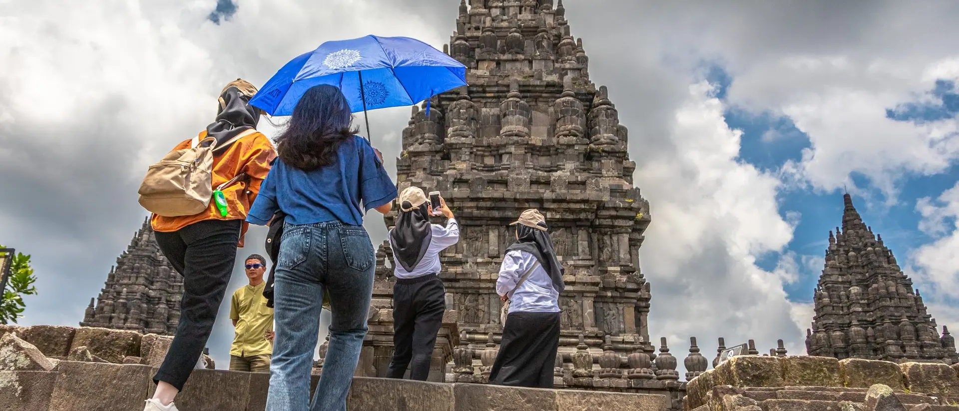 Visitors ascend stone steps toward ancient temples under a partly cloudy sky, some using umbrellas for shade.