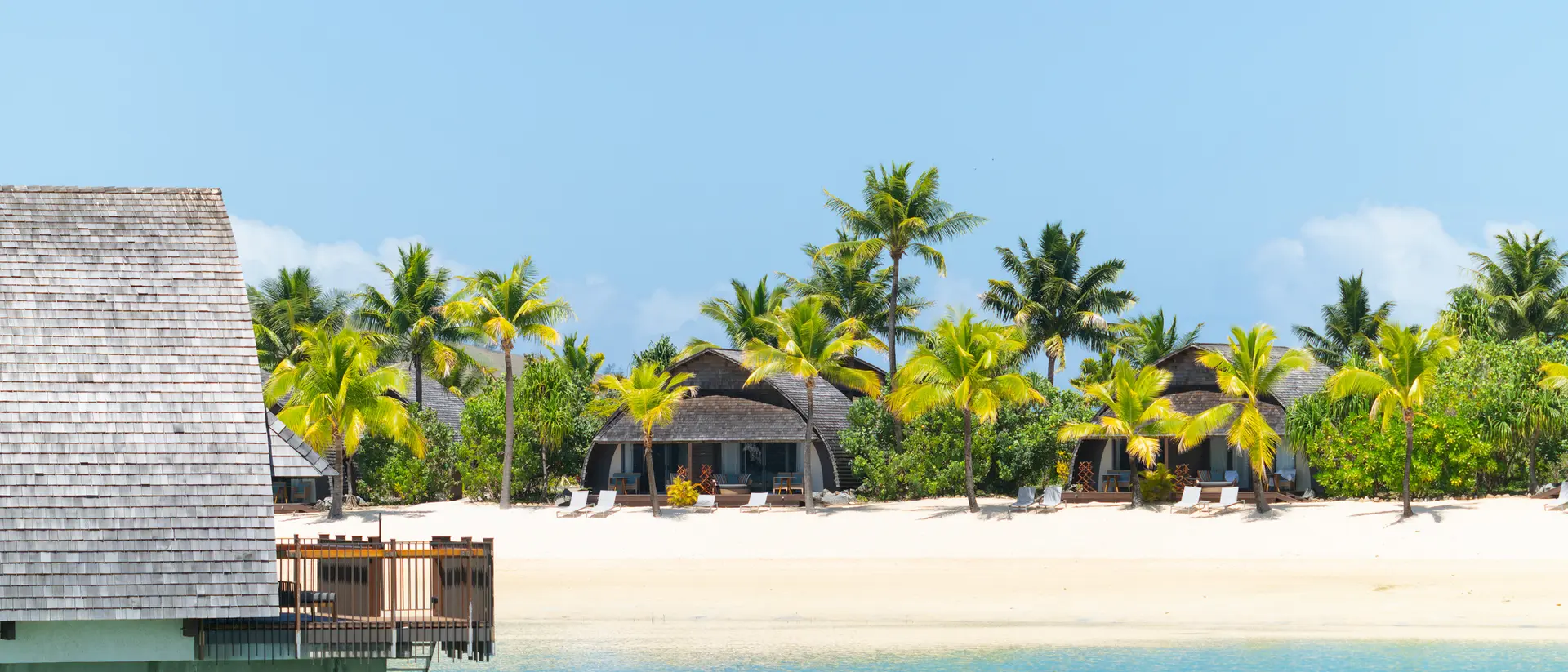 Tropical beach scene with palm trees, clear blue water, and thatched-roof huts along the shore under a bright sky.