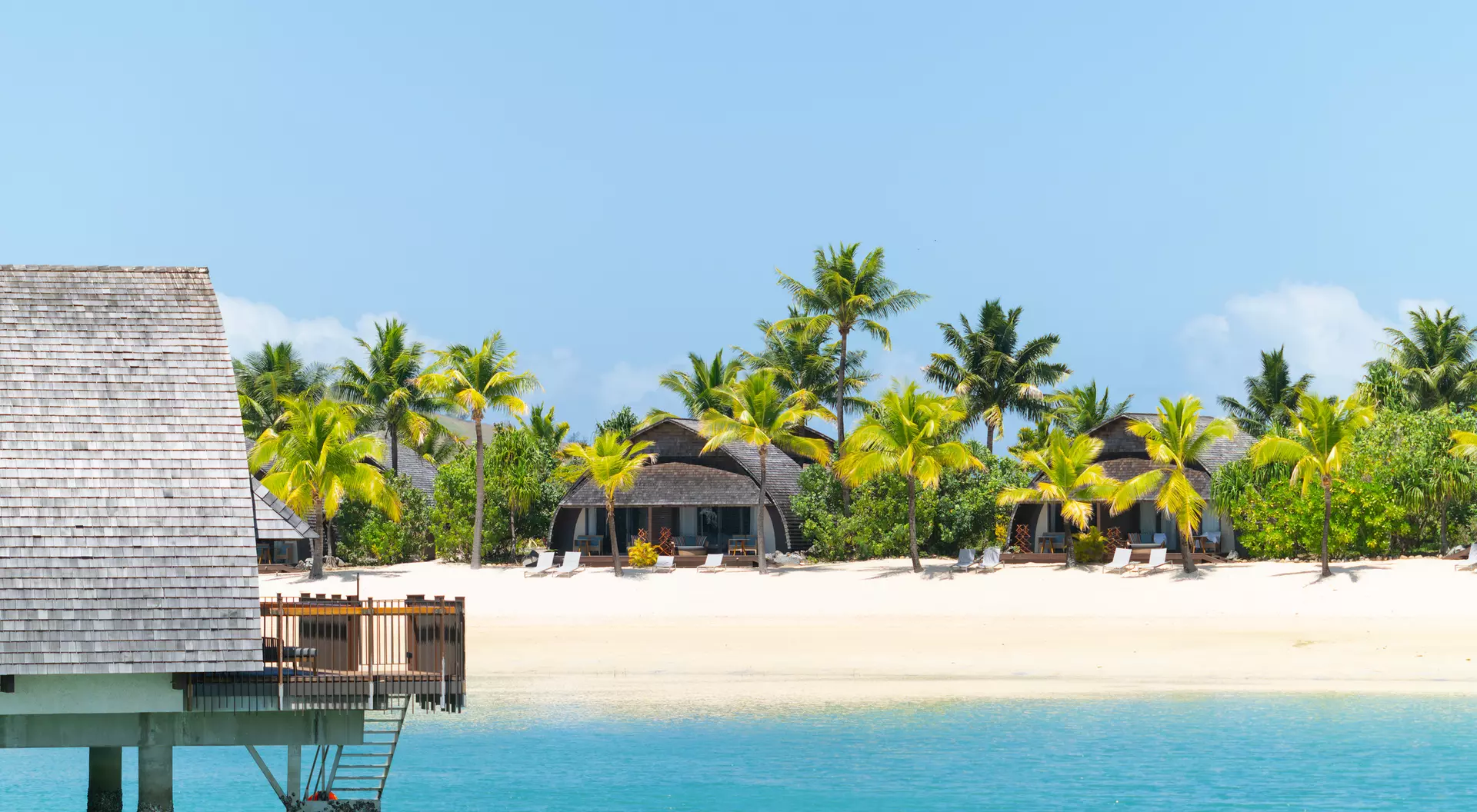 Tropical beach scene with palm trees, clear blue water, and thatched-roof huts along the shore under a bright sky.