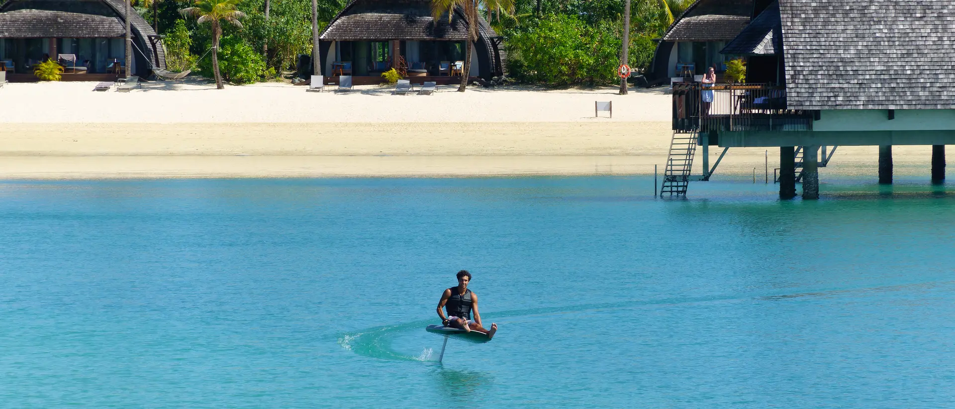 A person in a small boat glides across clear blue water near tropical huts and a sandy beach under a bright sky.