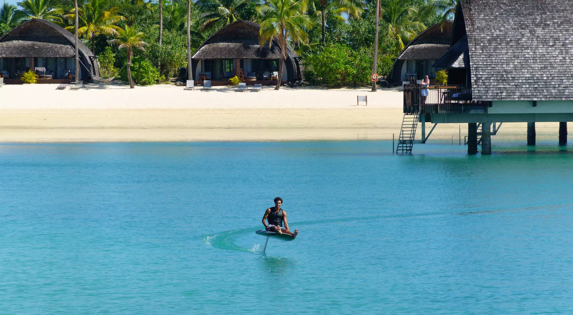 A person in a small boat glides across clear blue water near tropical huts and a sandy beach under a bright sky.