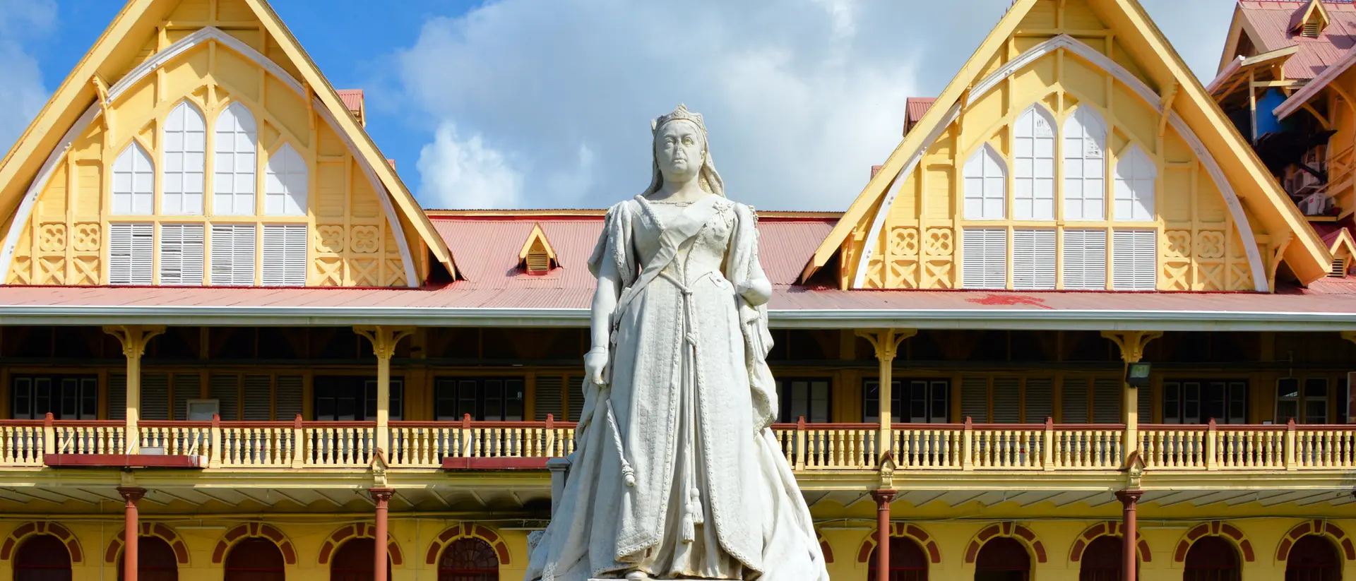 Statue of a woman in white dress in front of a historic yellow building with peaked roofs and blue sky.