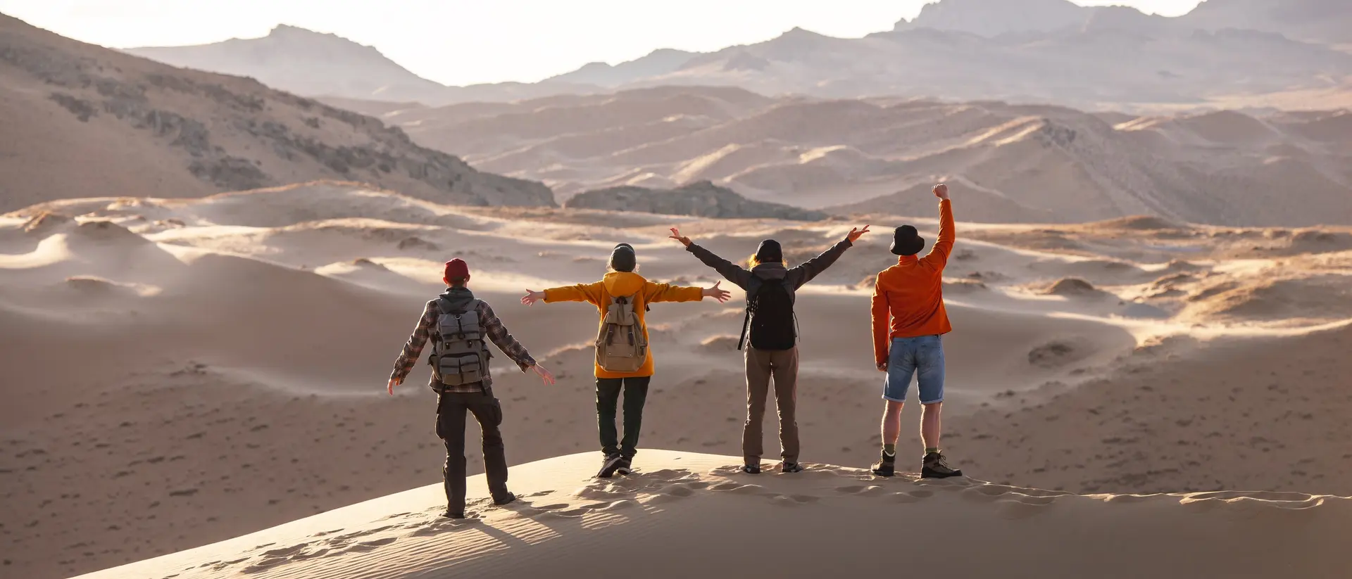 Four people stand on a sand dune, facing away and raising their arms, with vast desert landscape stretching behind them.
