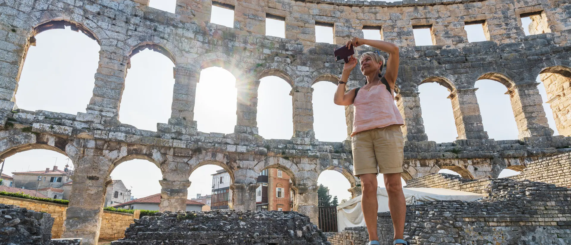 A person stands on rocks, photographing the ruins of a coliseum with sunlight streaming through the arches.