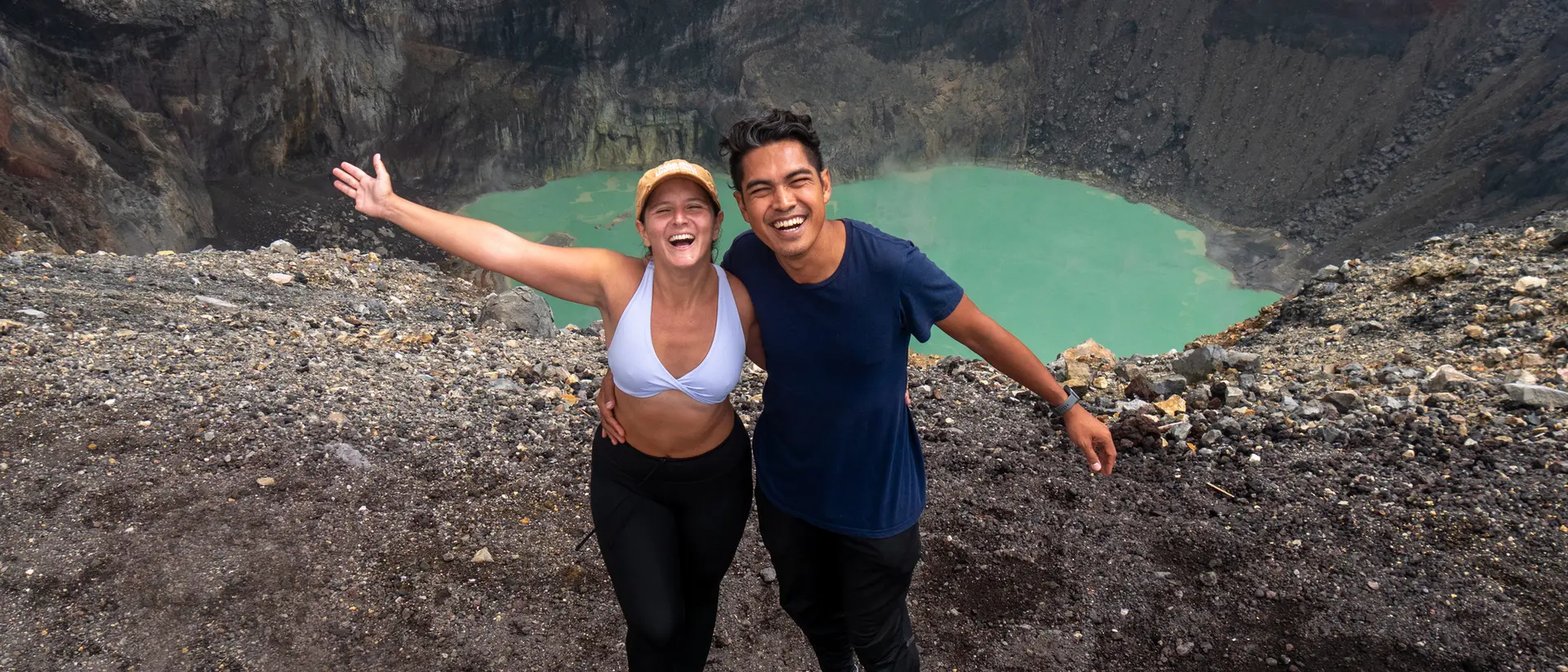 Two smiling people pose near a volcanic crater, with turquoise water visible below, against a dramatic rocky landscape.