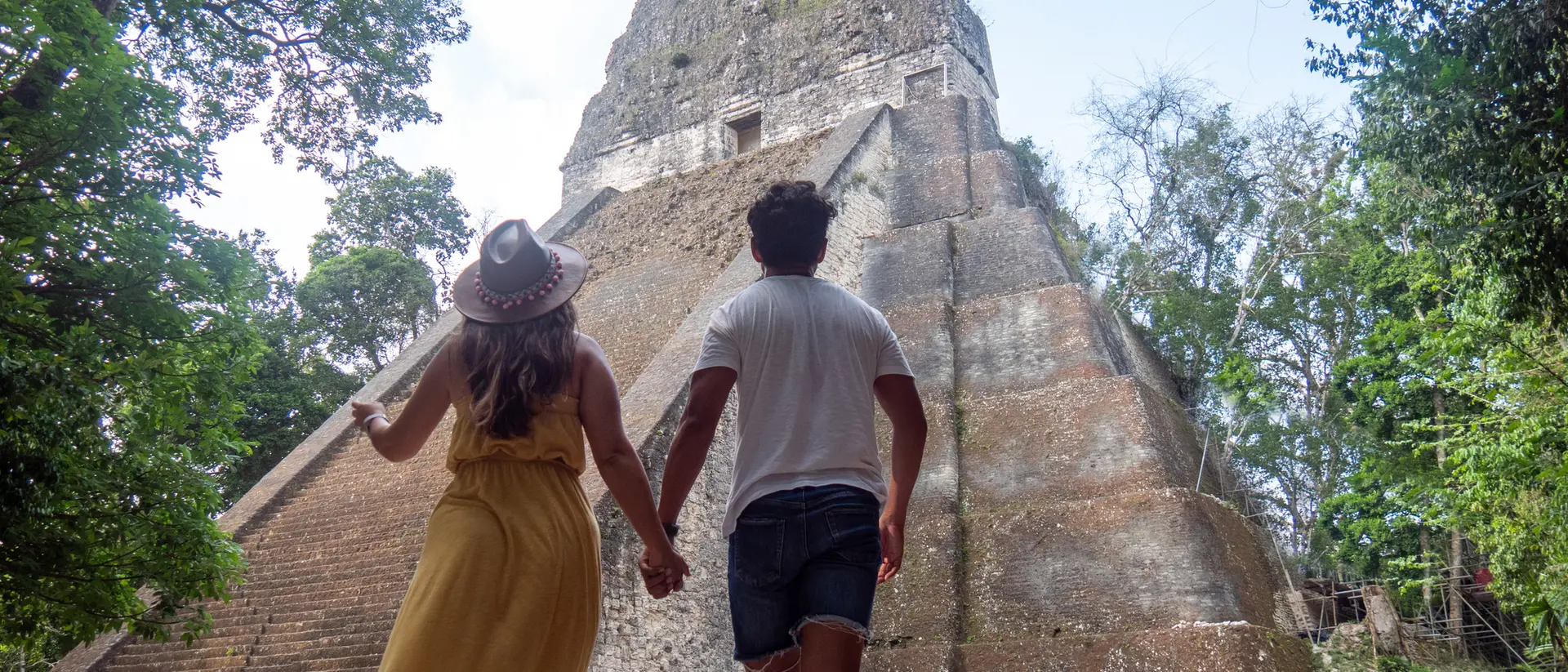 A couple holds hands walking towards a large ancient pyramid, surrounded by lush greenery under a bright sky.
