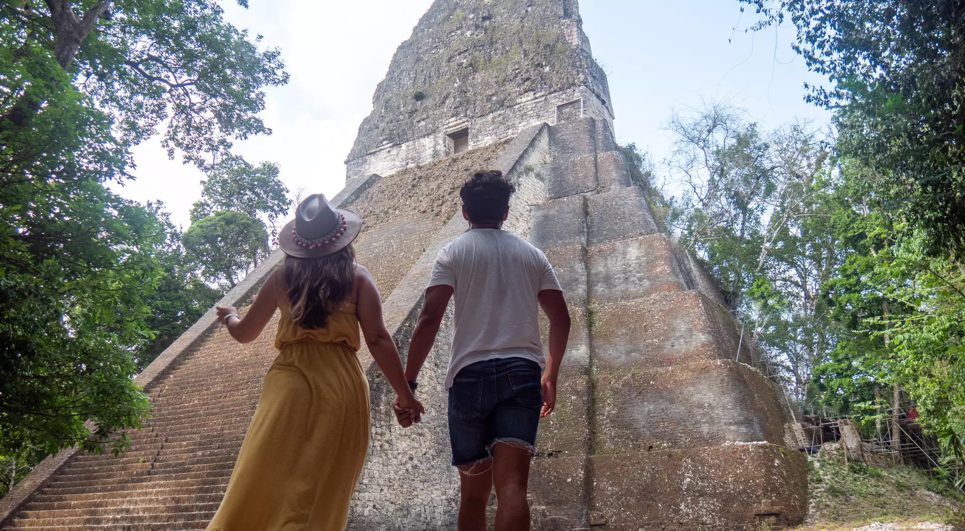 A couple holds hands walking towards a large ancient pyramid, surrounded by lush greenery under a bright sky.