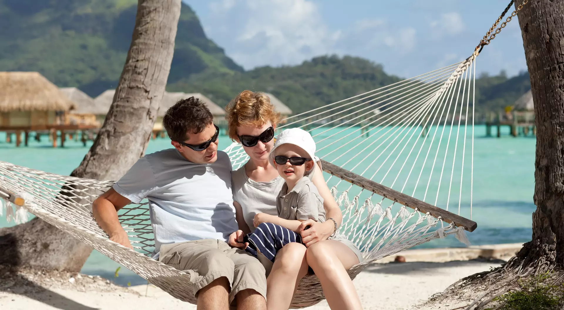 A family sits together on a hammock by the beach, enjoying the sun and surrounded by tropical scenery.