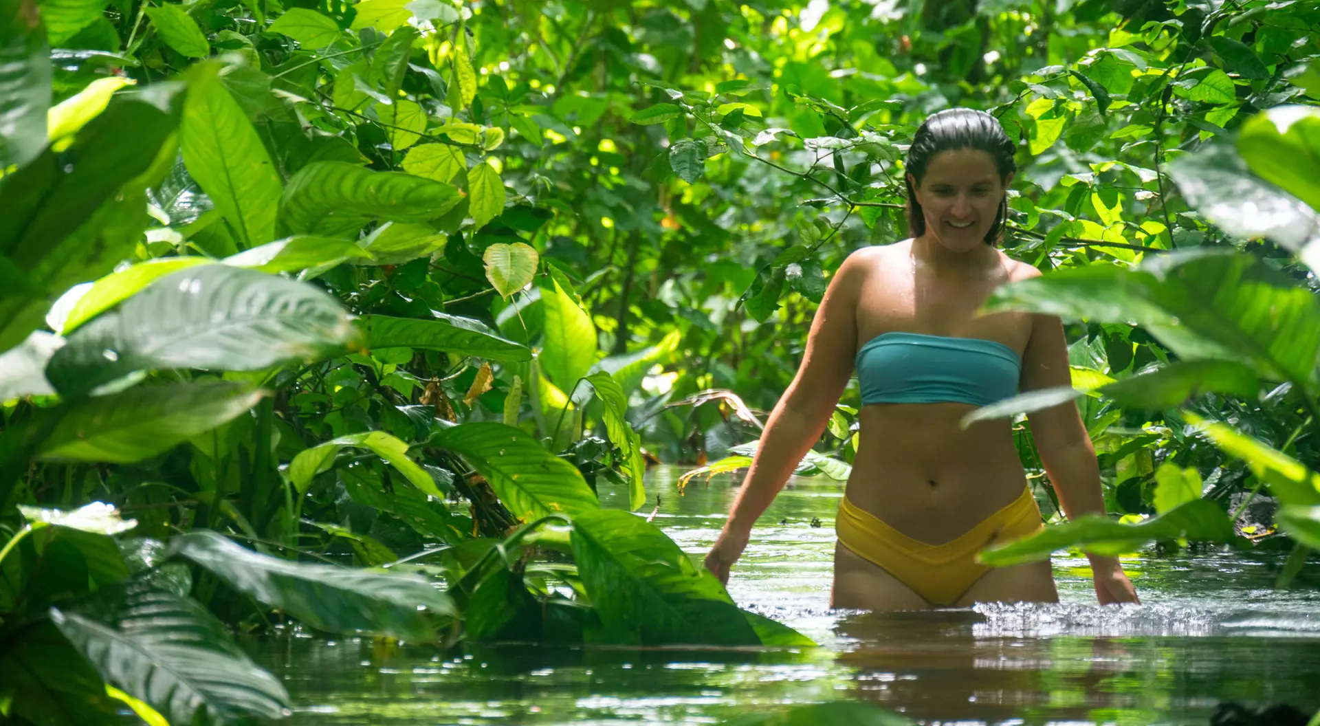 A woman wades through a lush, green jungle stream, surrounded by vibrant foliage and sunlight.