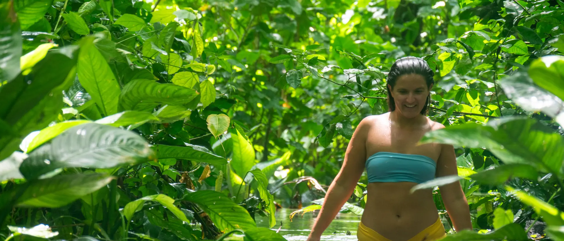 A woman wades through a lush, green jungle stream, surrounded by vibrant foliage and sunlight.