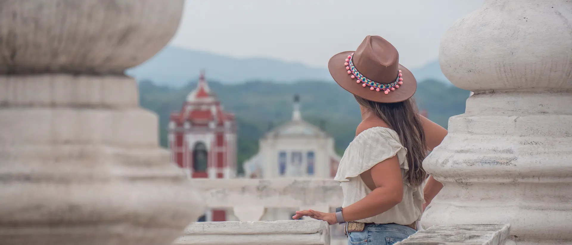 A woman in a tan hat stands on a terrace, gazing at colorful buildings and mountains in the distance.