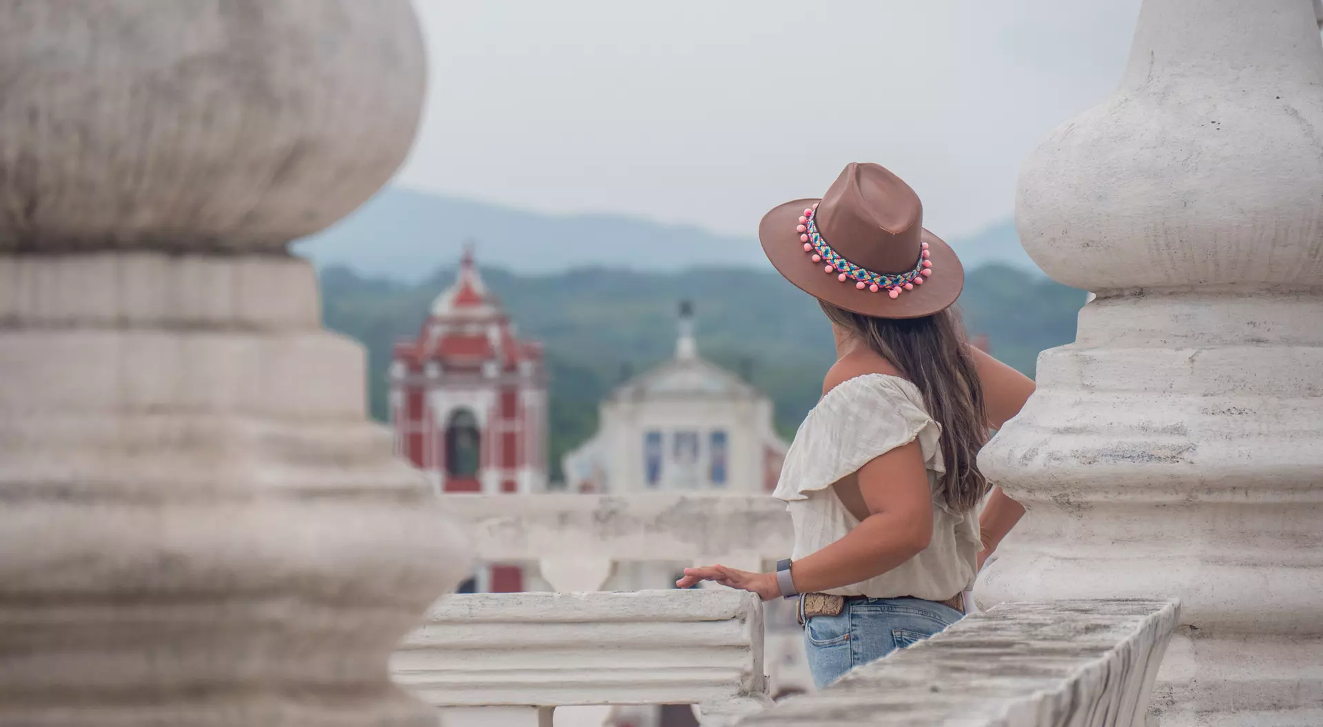 A woman in a tan hat stands on a terrace, gazing at colorful buildings and mountains in the distance.