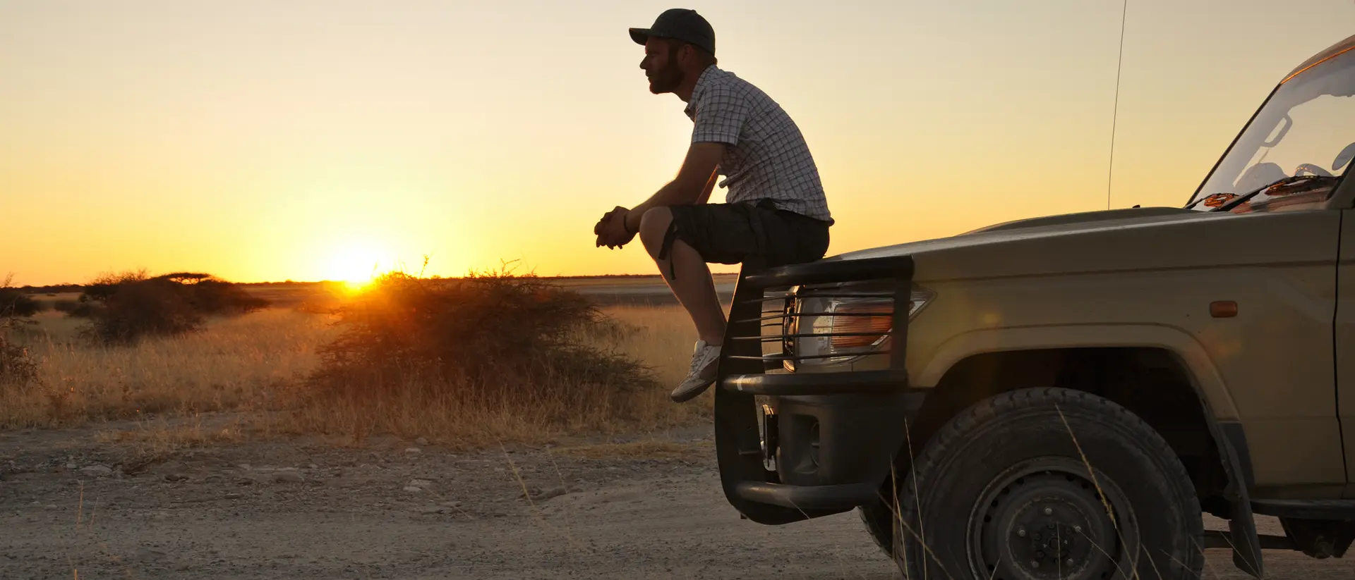 A person sitting on the front of a vehicle, overlooking a sunset in a grassy landscape.