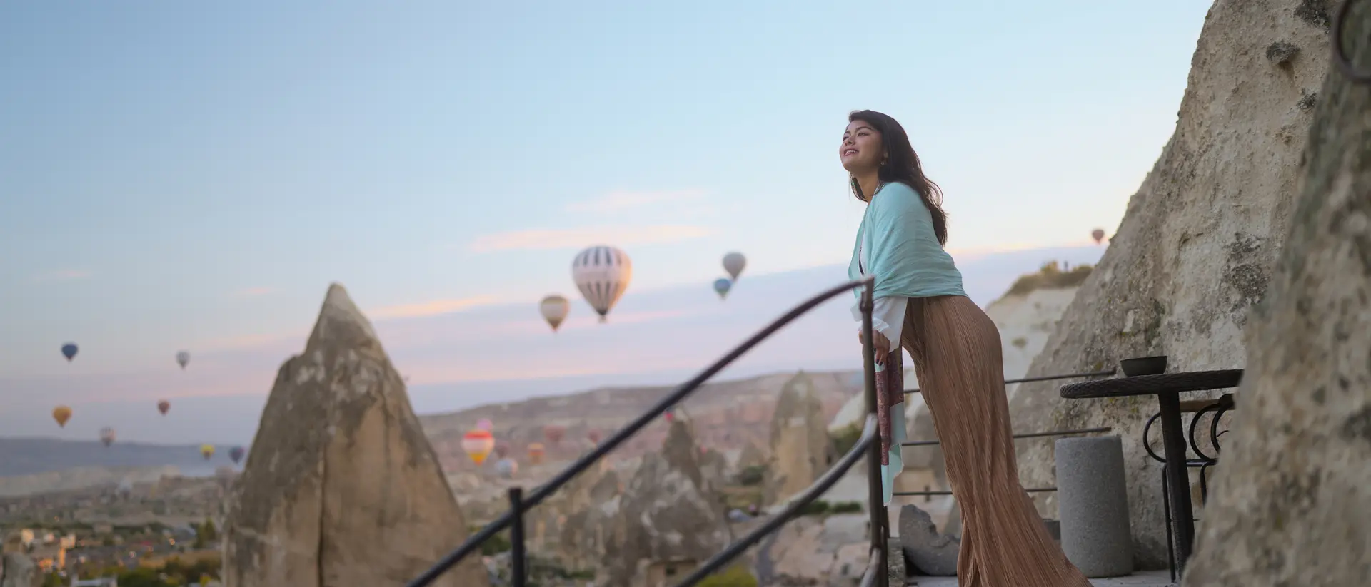 A woman stands on a balcony overlooking a valley with hot air balloons in the sky at sunset.