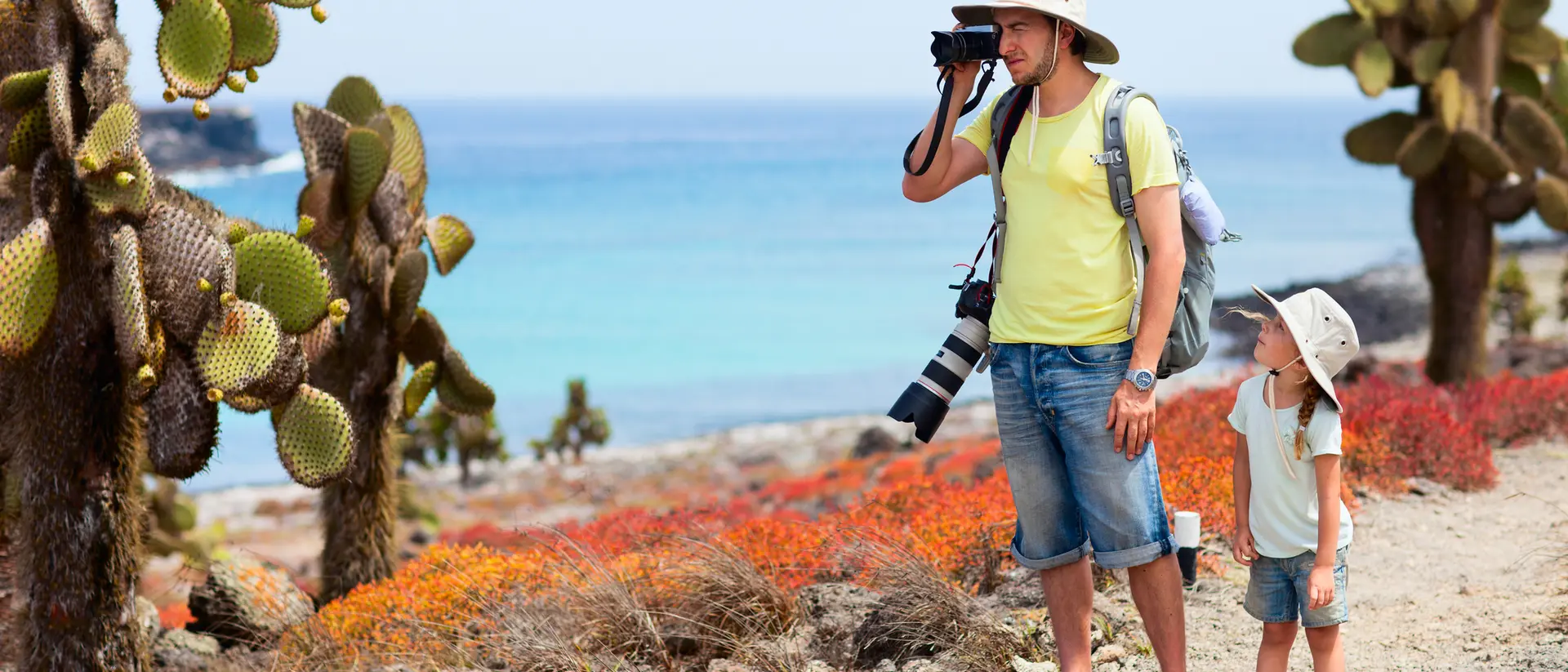 A man taking photos of cacti with a camera, accompanied by a child wearing a hat near a blue ocean.