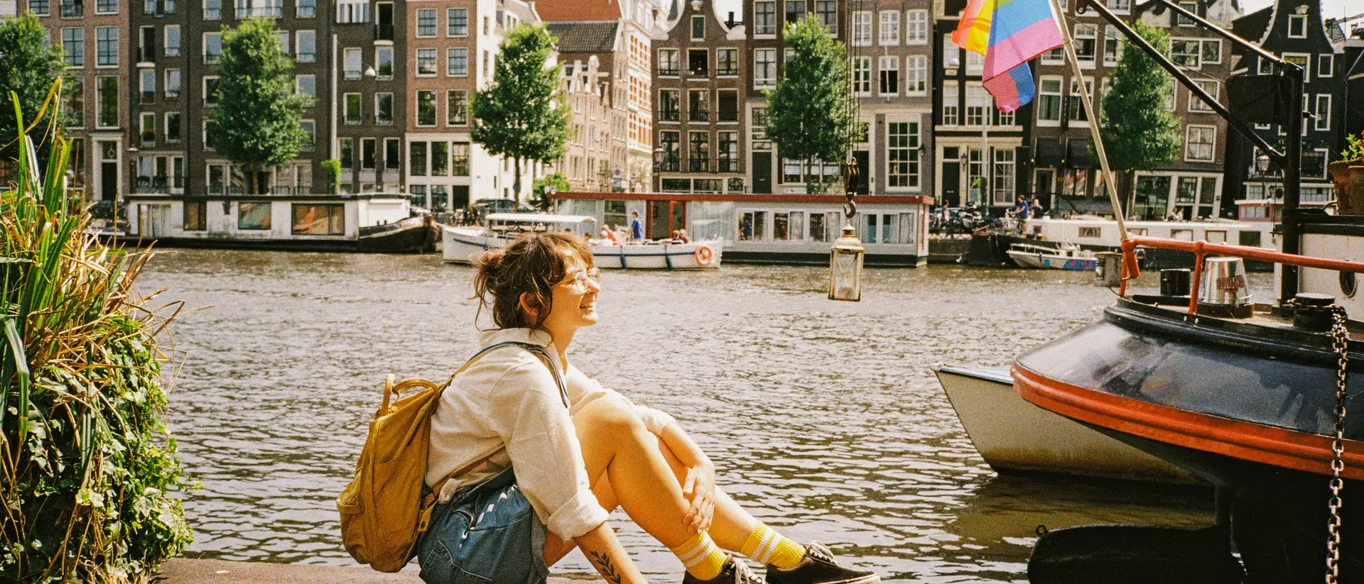 Person sitting by a canal in Amsterdam with colorful buildings and a rainbow flag in the background on a sunny day.