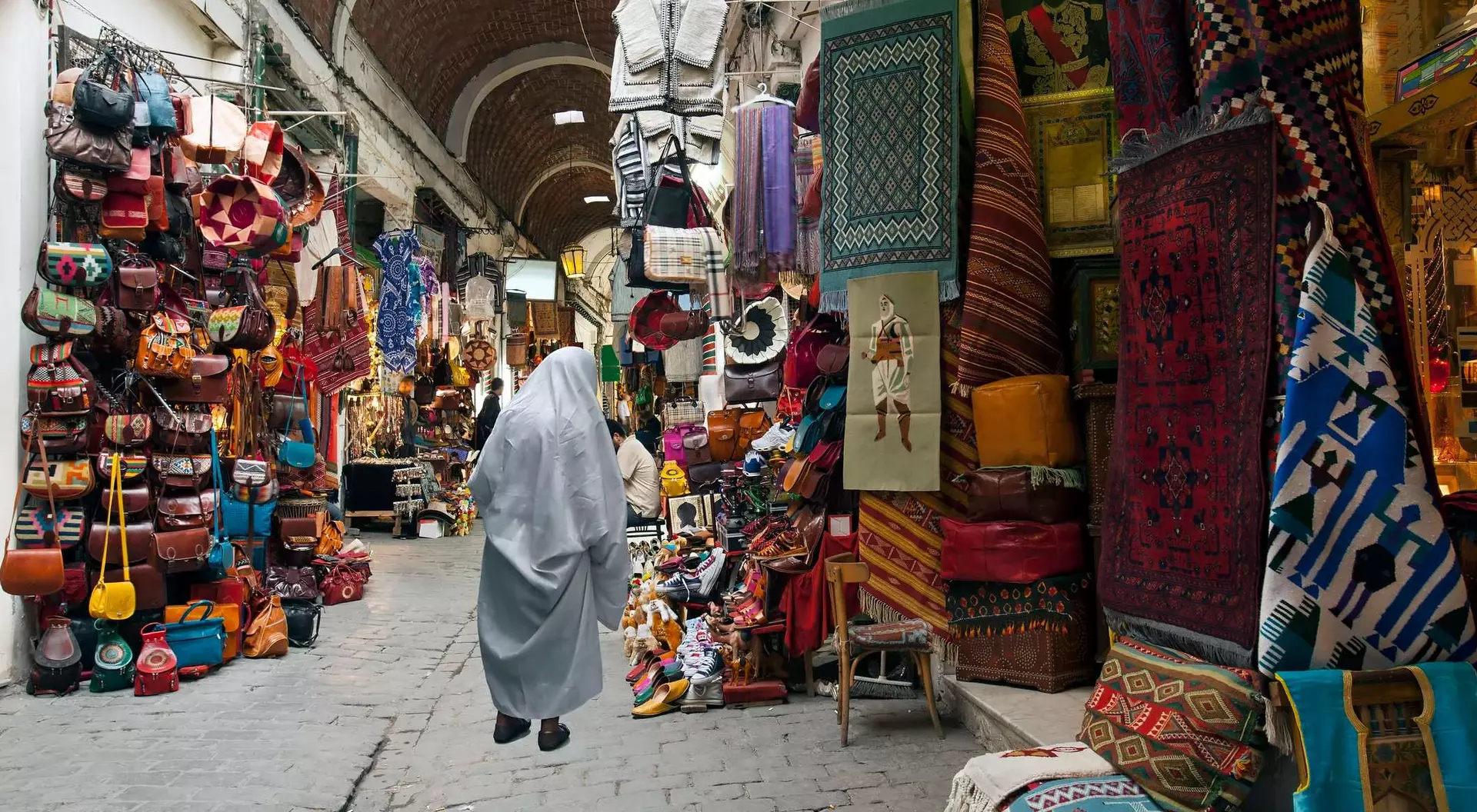 A bustling market with vibrant textiles, bags, and crafts, featuring a person in a gray cloak walking through the narrow path.