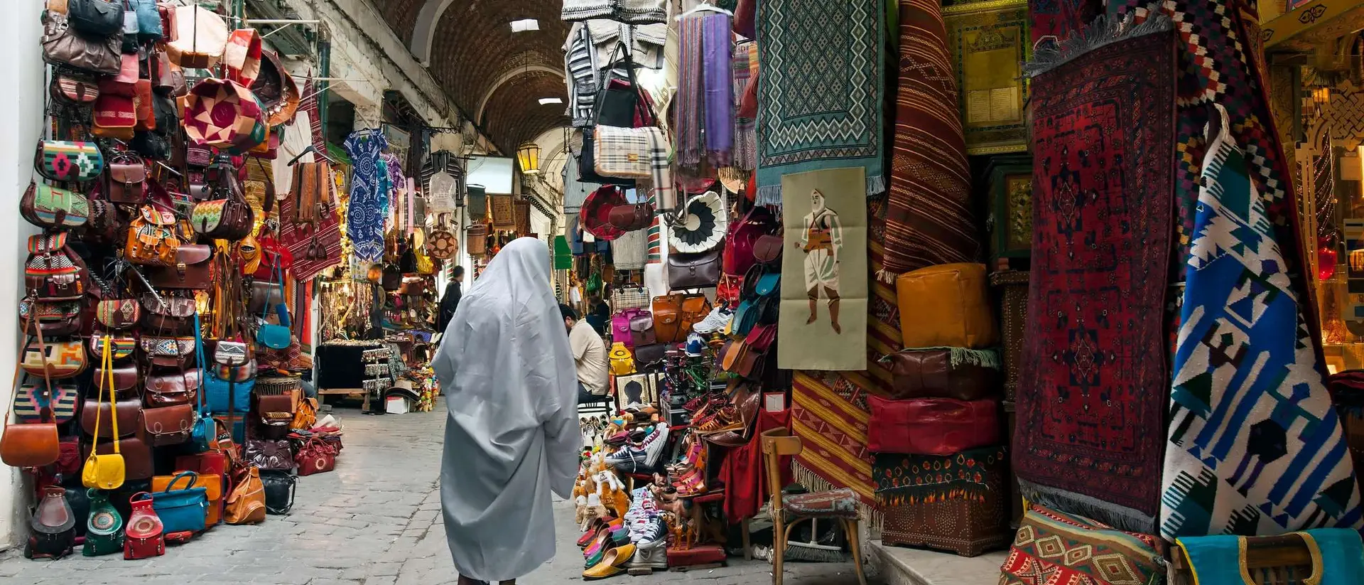 A bustling market with vibrant textiles, bags, and crafts, featuring a person in a gray cloak walking through the narrow path.