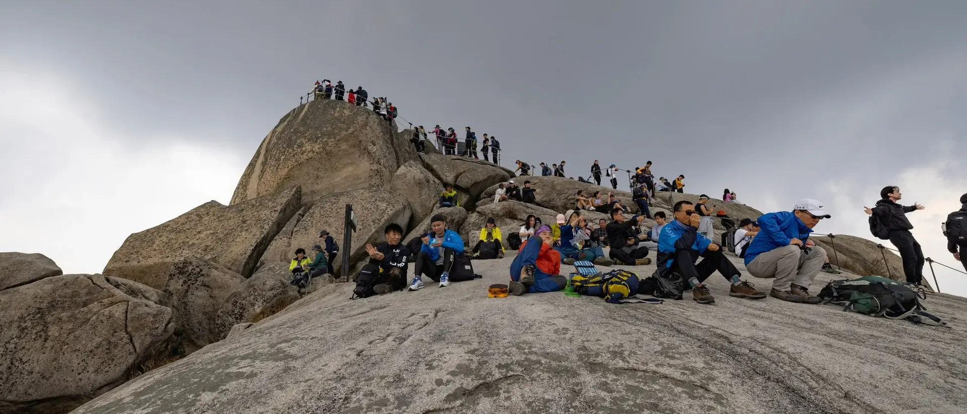 A group of hikers resting on a rocky summit under cloudy skies, with more climbers in the background.