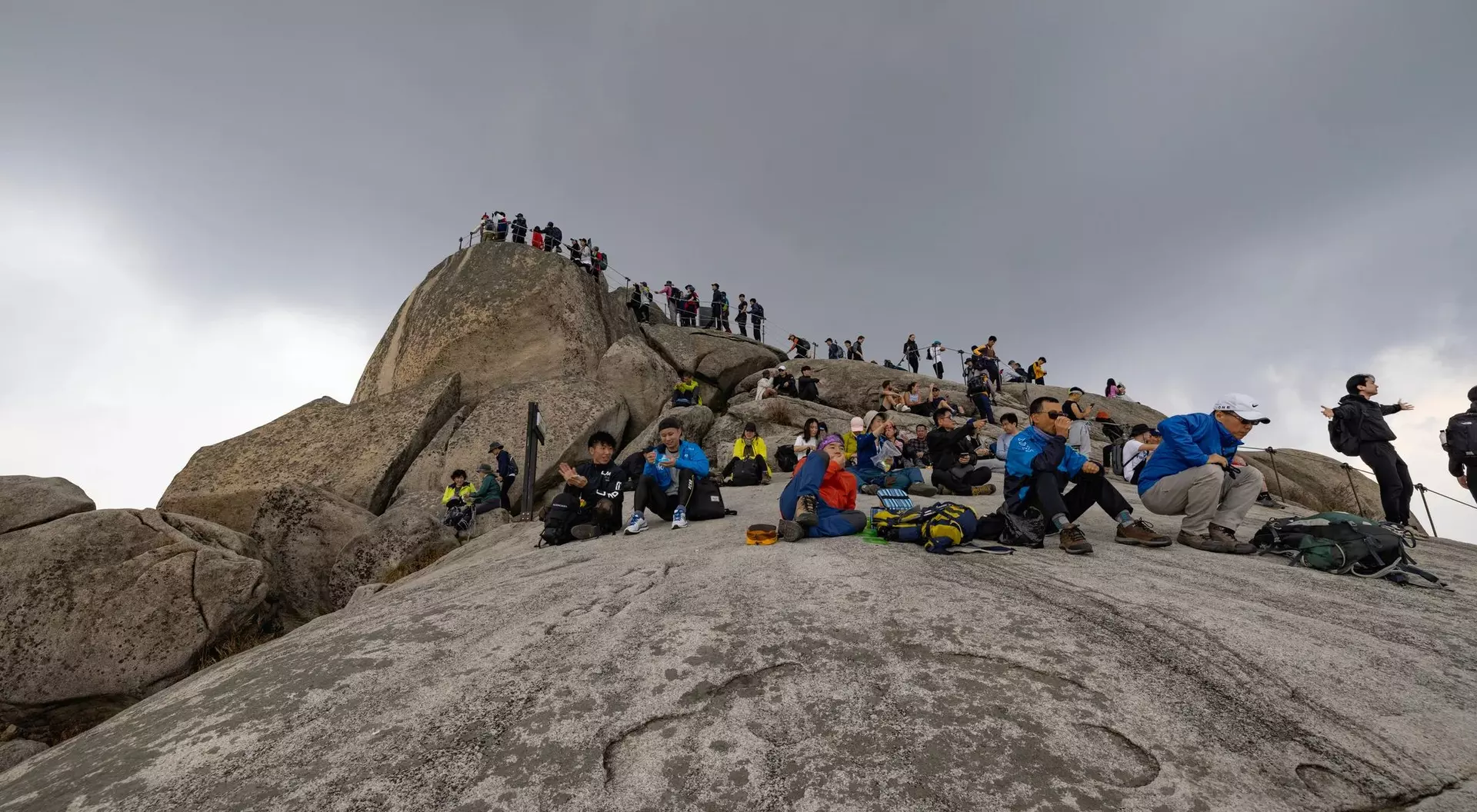 A group of hikers resting on a rocky summit under cloudy skies, with more climbers in the background.