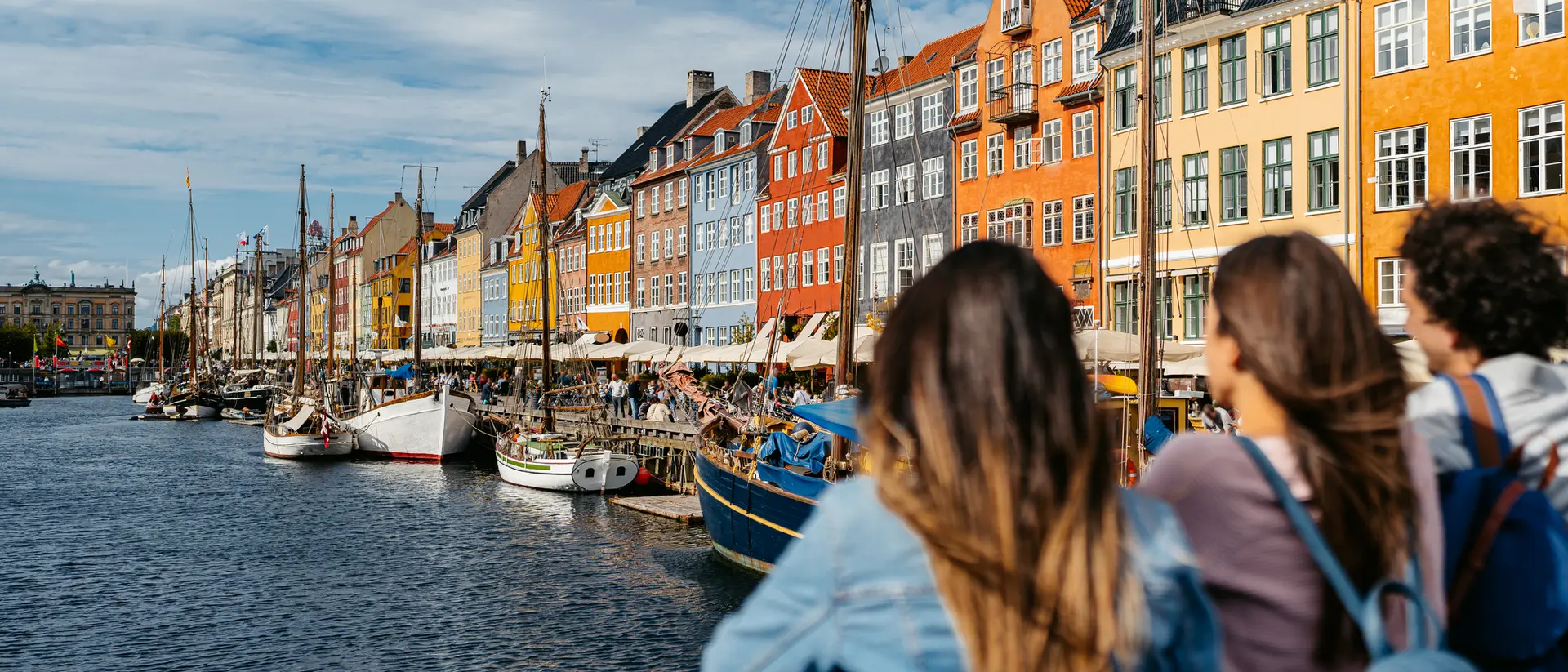 Three people stand by a canal, gazing at colorful buildings and boats in a scenic waterfront view.