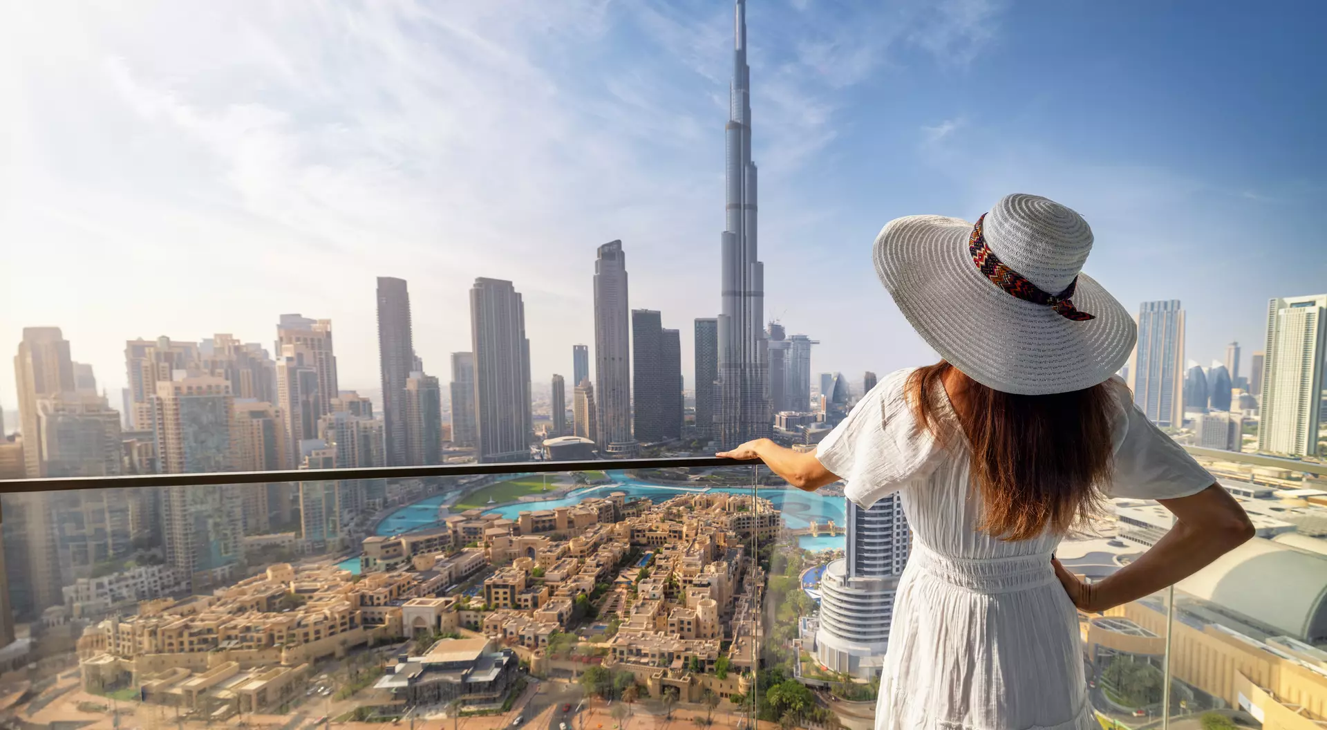 A woman in a white dress and hat gazes at a city skyline featuring the Burj Khalifa in the background.