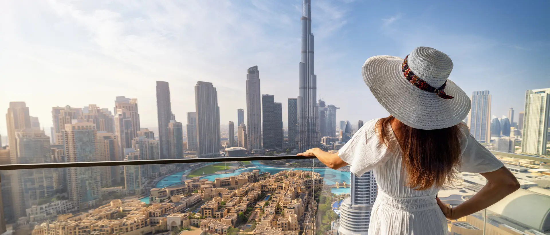 A woman in a white dress and hat gazes at a city skyline featuring the Burj Khalifa in the background.