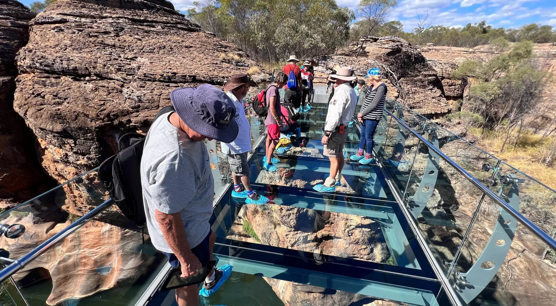 A group of people standing on a glass bridge surrounded by rocky terrain and blue sky.