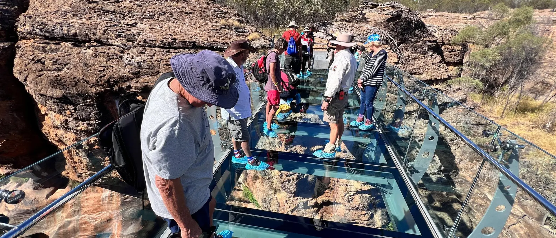 A group of people standing on a glass bridge surrounded by rocky terrain and blue sky.