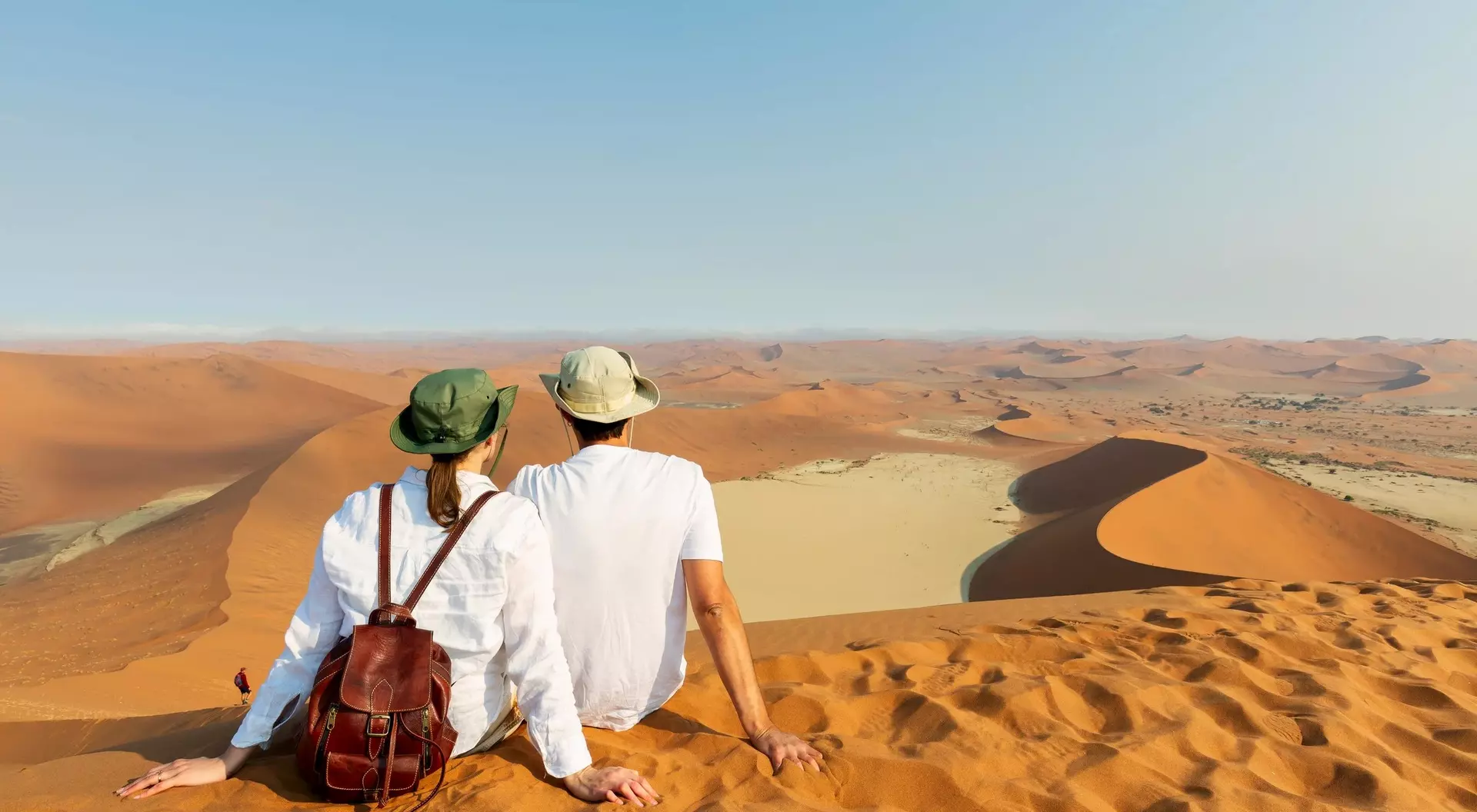 A couple sits on a sandy dune, overlooking vast desert landscapes under a clear sky.
