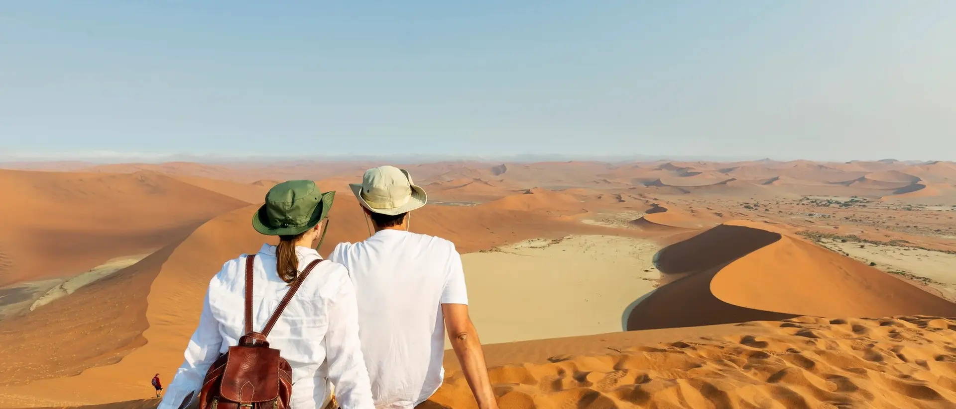 A couple sits on a sandy dune, overlooking vast desert landscapes under a clear sky.