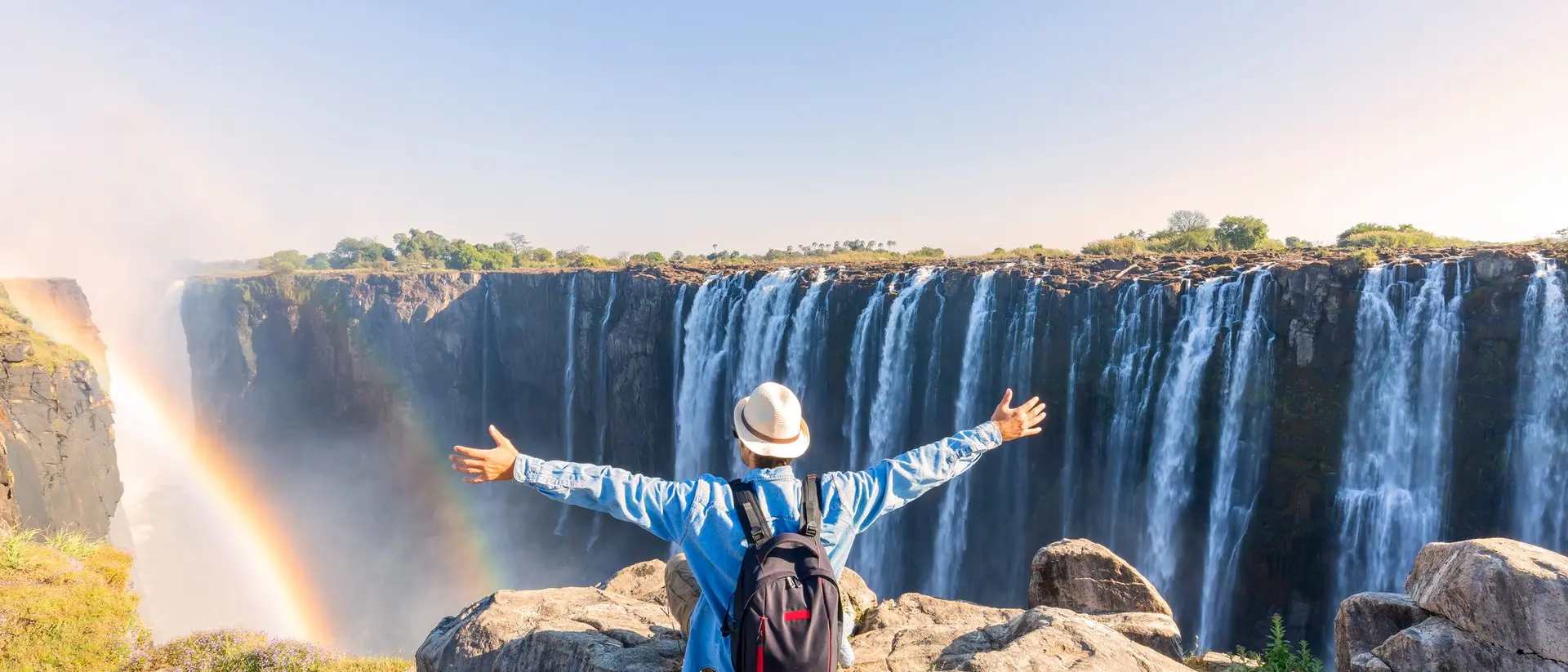 Person with a backpack and hat standing on rocks, arms outstretched, overlooking a majestic waterfall with a rainbow.