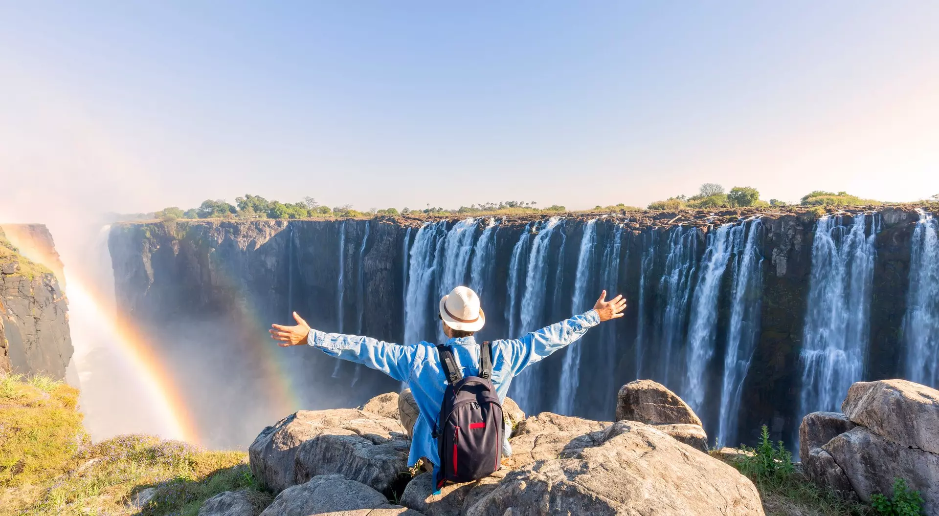 Person with a backpack and hat standing on rocks, arms outstretched, overlooking a majestic waterfall with a rainbow.