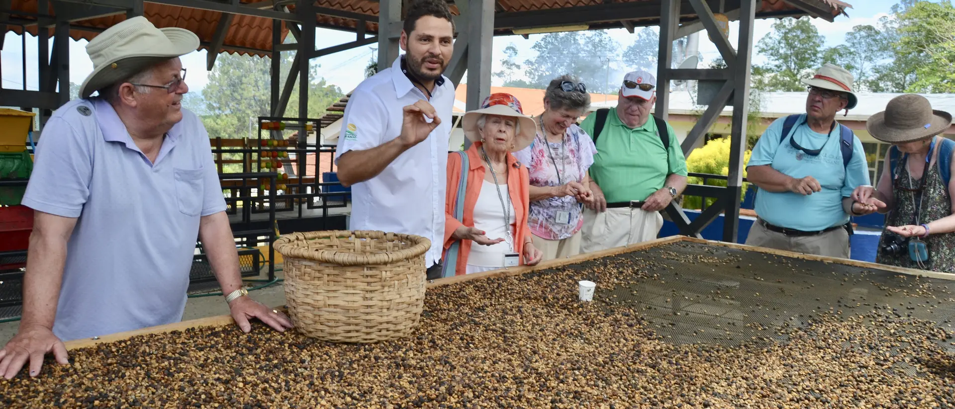 A group of tourists listens to a guide near a large table covered with beans, under a covered outdoor area.