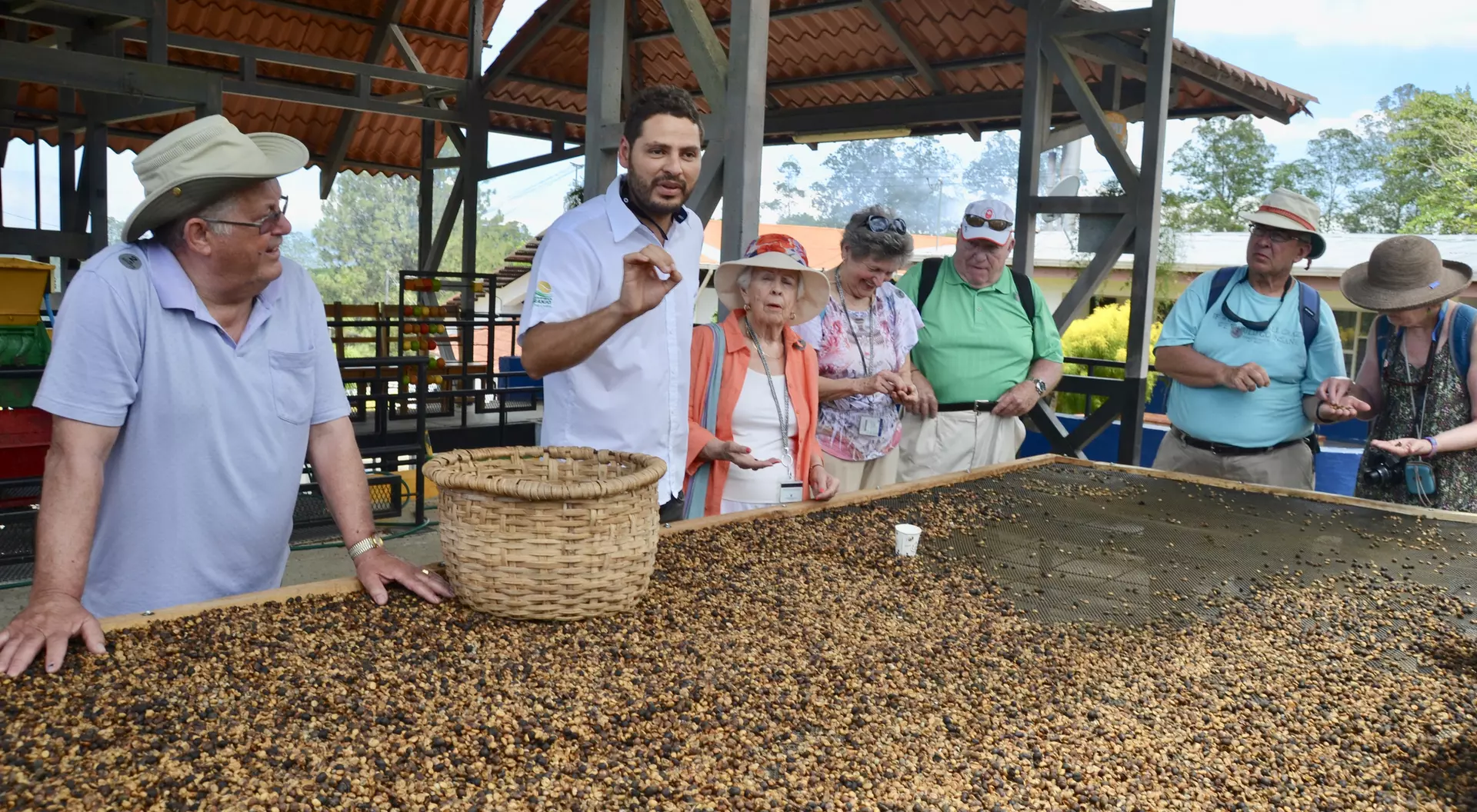 A group of tourists listens to a guide near a large table covered with beans, under a covered outdoor area.