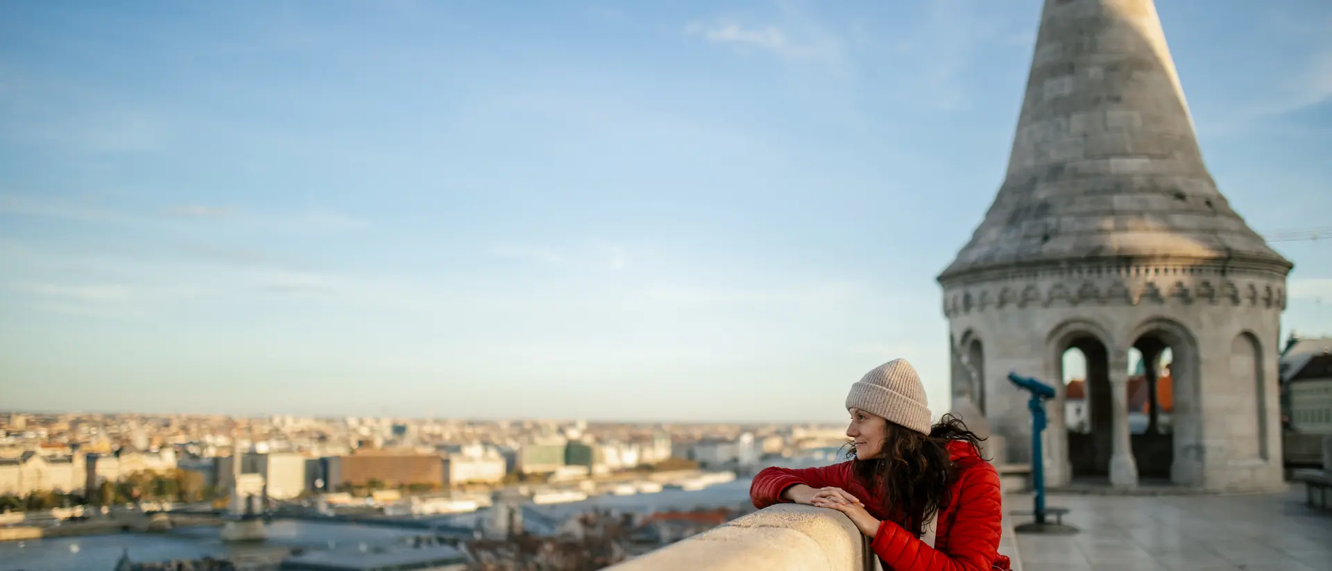 A person in a red jacket and a hat leans on a stone railing, overlooking a cityscape with a tall tower in the background.