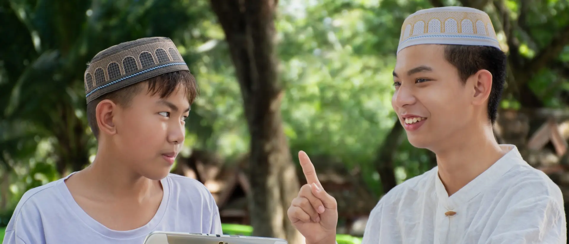 Two young men wearing traditional hats engage in conversation outdoors, one holding a tablet and listening intently.