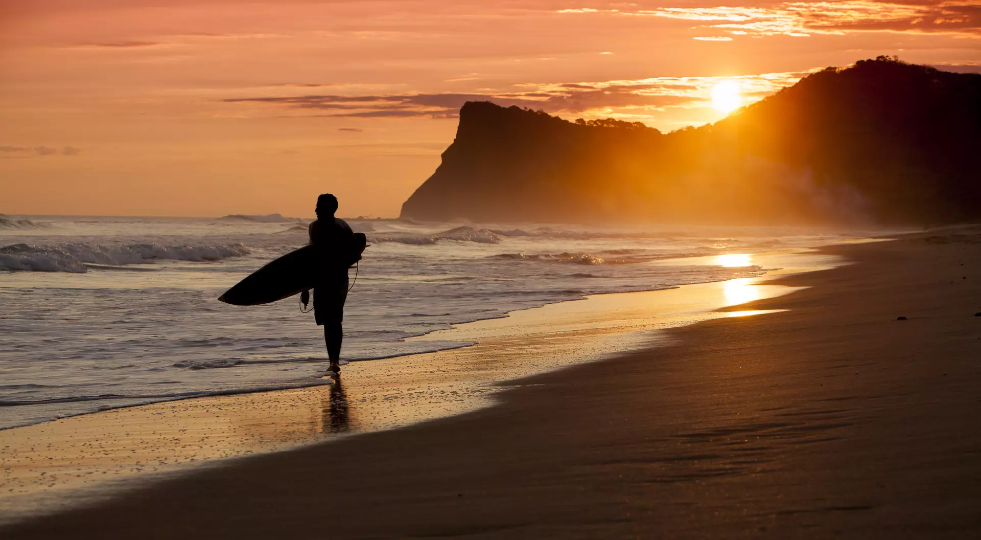 Silhouette of a person walking on the beach with a surfboard at sunset, waves gently lapping at the shore.