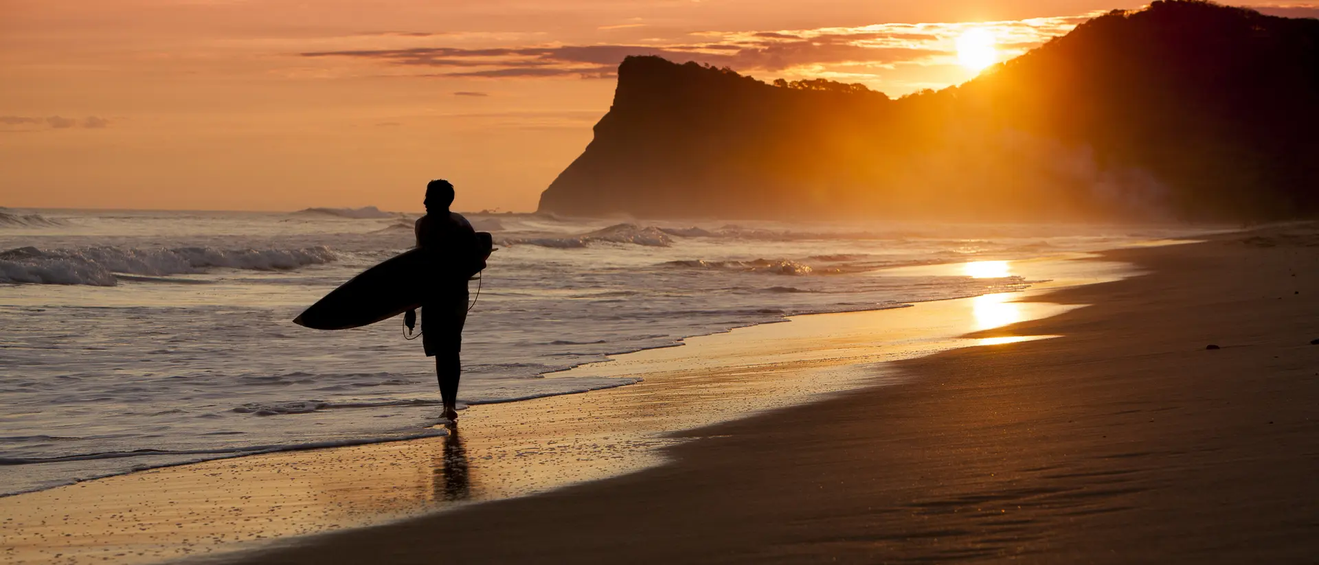 Silhouette of a person walking on the beach with a surfboard at sunset, waves gently lapping at the shore.