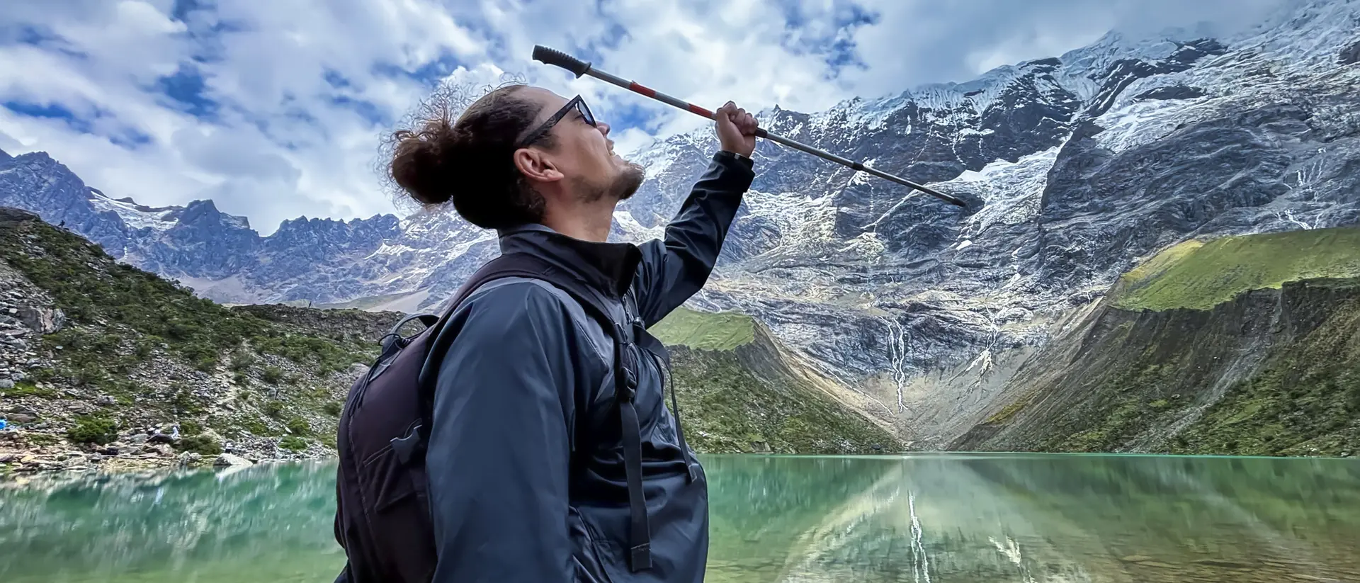 Person with a backpack raises a walking stick in front of a tranquil lake surrounded by mountains and a cloudy sky.
