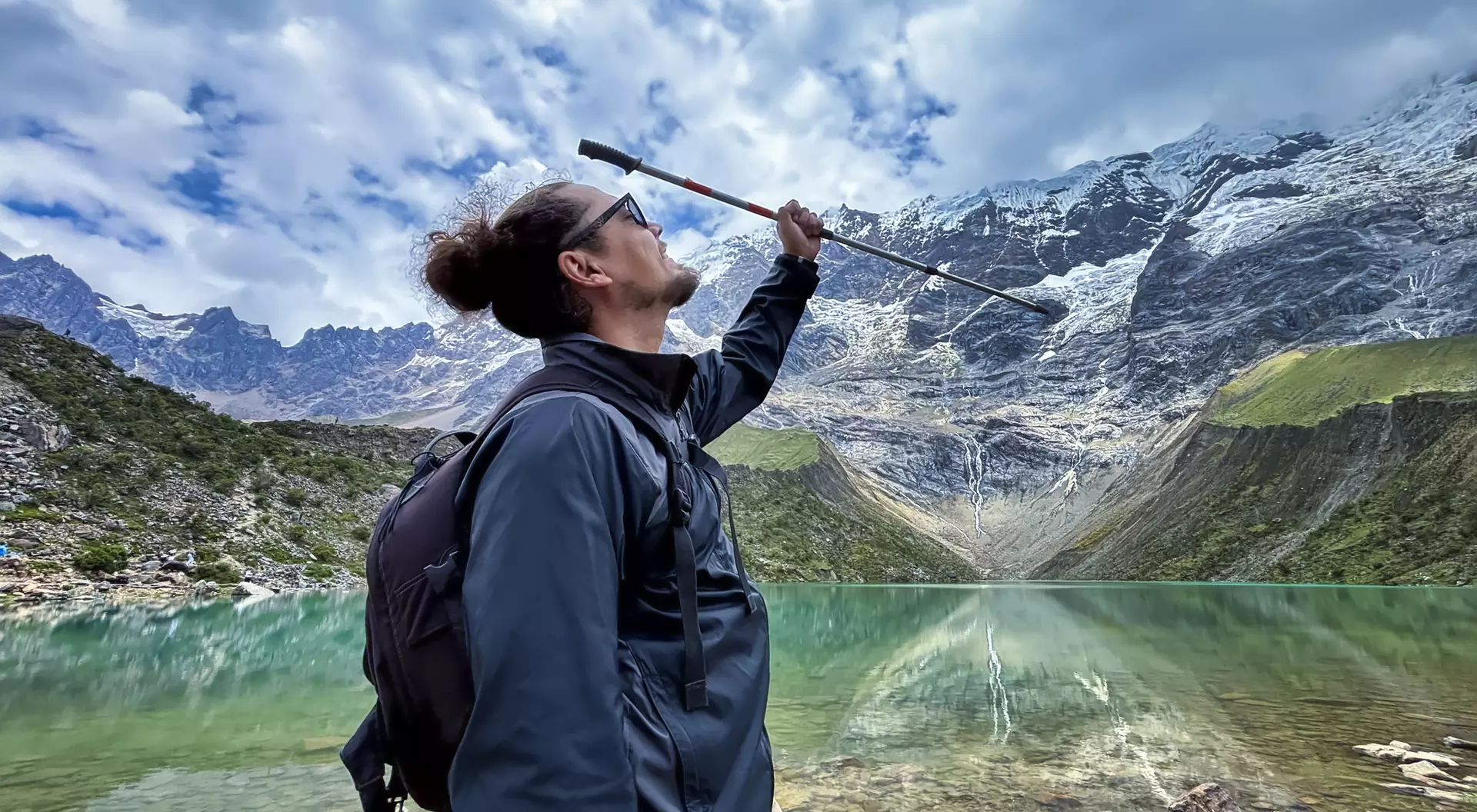 Person with a backpack raises a walking stick in front of a tranquil lake surrounded by mountains and a cloudy sky.