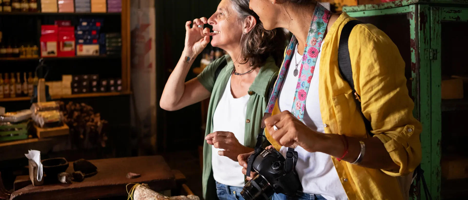 Two women enjoy a moment in a market, surrounded by various cured meats and goods, one holding a camera.